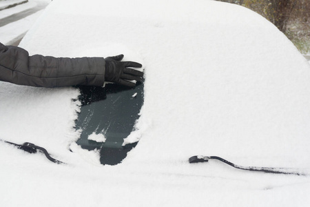 一个戴着手套的妇女在清扫汽车上的积雪。积雪覆盖的汽车挡风玻璃。冬天下雪时停着的车被雪覆盖。
