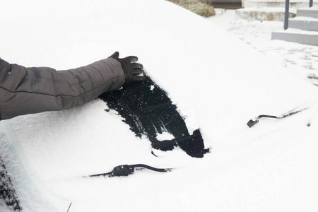 一个戴着手套的妇女在清扫汽车上的积雪。积雪覆盖的汽车挡风玻璃。冬天下雪时停着的车被雪覆盖。