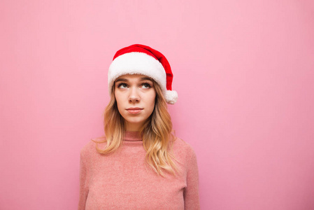 可爱的女士穿着粉红色的毛衣，戴着圣诞老人的帽子，在粉红色的背景下，抬起头来，面带严肃。戴着圣诞帽的女孩看着空旷的空间。Xmas
