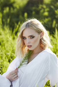 Girl in a long white dress sits on the grass in a field. Blonde 