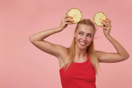 美丽的年轻的金发女子，笑容愉快地看着一边，在粉红色的背景上摆出新鲜菠萝片的姿势
