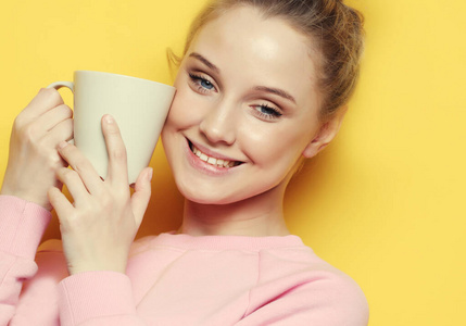年轻的金发女子拿着白杯子喝茶或咖啡，生活方式和饮食观念。闭合。黄色背景。