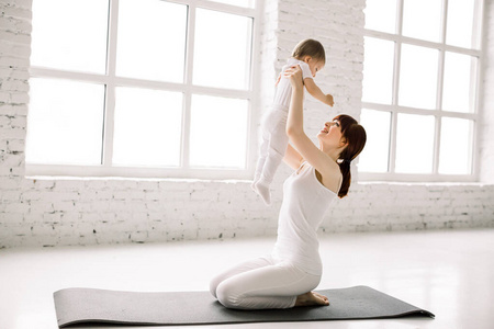 一位母亲和婴儿在白色的墙壁和大窗户的背景下做体操,瑜伽练习.