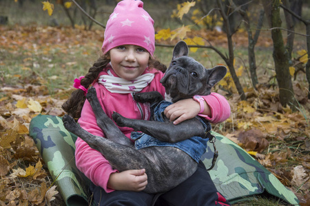 In the autumn park, a little girl holds a French bulldog in her 