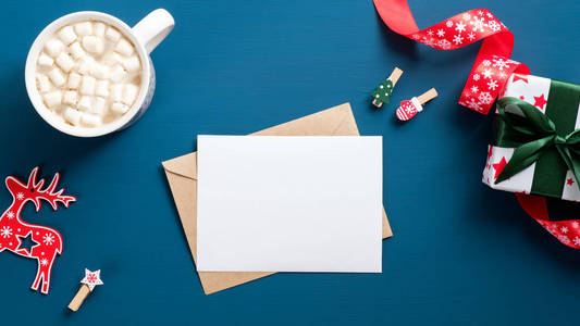 蓝色背景上的圣诞信。平铺空白纸卡，工艺纸信封，咖啡杯，圣诞装饰品，礼品盒。给圣诞老人写信的概念