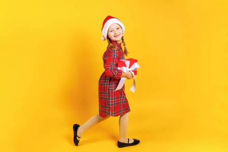 戴圣诞老人帽的快乐小女孩偷偷带着圣诞礼物。一个在黄色背景下发育完全的孩子