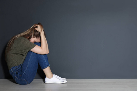 坐在灰色墙壁附近地板上的沮丧的年轻女人.文本空间照片