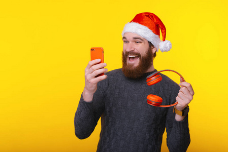兴奋的男人戴着圣诞老人帽，看着智能手机，手里拿着红色无线耳机