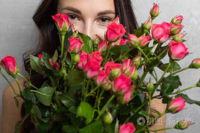 在灰色背景下一位身着夏装手持一束玫瑰花看着相机的迷人年轻女子的