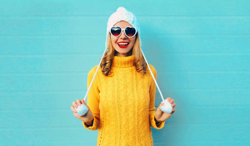 冬季肖像快乐微笑的年轻女子穿着黄色针织毛衣和白色帽子，蓝色墙壁背景上戴着心形太阳镜