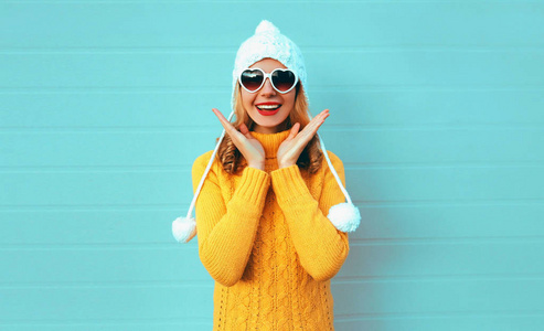 冬季肖像快乐惊喜年轻女子穿着黄色针织毛衣和白色帽子，蓝色墙壁背景上戴着心形太阳镜