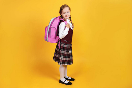 一个穿制服的女学生背着背包站在黄色背景上。可爱的小女孩微笑着看着镜头