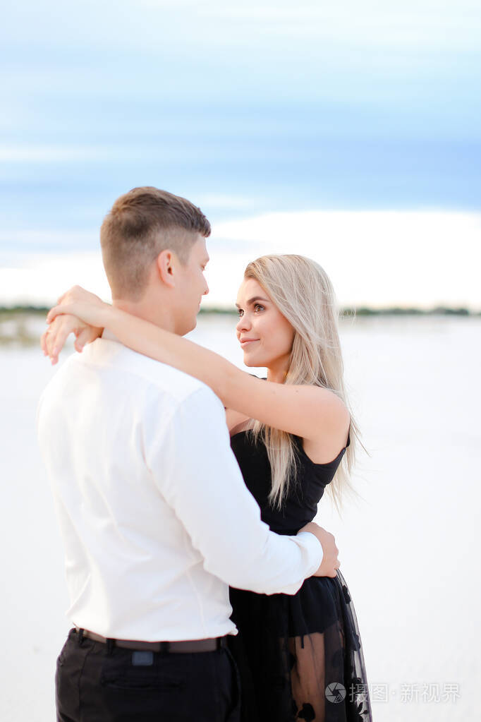 穿着黑色连衣裙的年轻金发女人在冬天的田野背景下拥抱着男人