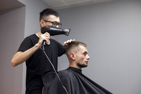 帅哥在理发店理发,哈萨克理发师靠着灰色的墙给客户吹风,理发师剪短发