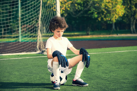 夏季足球锦标赛。情绪游戏的乐趣。坐在球上的年轻进球。球场上穿着足球运动服的男孩守门员。运动理念。