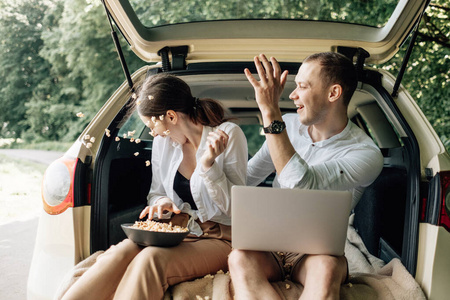 年轻幸福的情侣穿着同样的白色T恤坐在汽车后备箱里，在路边放着笔记本电脑和爆米花，周末出城，度假和公路旅行的概念
