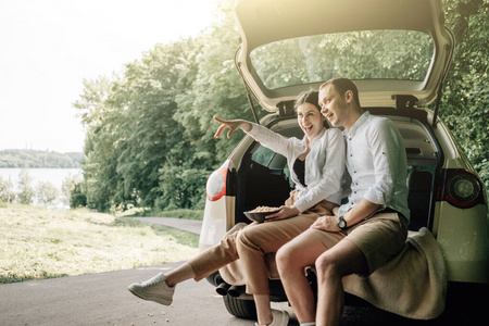 年轻幸福的情侣穿着同样的白色T恤坐在汽车后备箱里，路边放着爆米花，周末出城，度假和公路旅行的概念