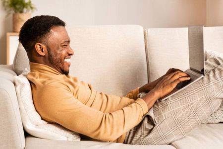 快乐的非洲裔美国人躺在沙发上用笔记本电脑