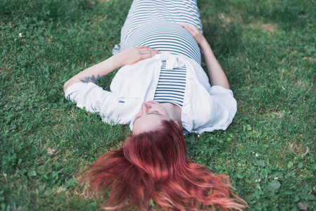 夏天，怀孕的白人女孩穿着一件浅条纹连衣裙躺在绿色的草地上。怀孕的女孩。夏天公园里怀孕的女孩。