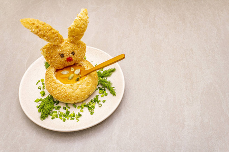 复活节兔子儿童食品概念。温热南瓜奶油汤在新鲜的面包卷，香草和南瓜子在盘中。石头背景，复制空间。