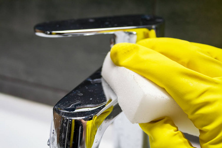 一只手戴着黄色橡胶手套，用白色三聚氰胺海绵擦拭肮脏的镀铬金属水龙头。在现代化的浴室里打扫卫生。选择性聚焦。特写视图