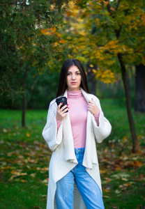 年轻的女性穿着白色开衫在秋天的公园里喝着黑咖啡