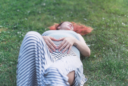 夏天，怀孕的白人女孩穿着一件浅条纹连衣裙躺在绿色的草地上。怀孕的女孩。夏天公园里怀孕的女孩。