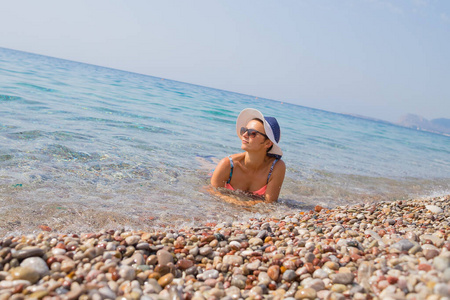 性感的女孩穿着比基尼和帽子，享受在卵石水海滩。漂亮女人在空荡荡的卵石沙滩上放松保留。好吧身材匀称的比基尼坐在水里，欣赏迷人