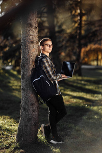 在公园里，穿着休闲装的聪明的男学生拿着笔记本或笔记本电脑站在树前看向别处。