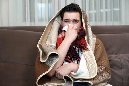 一个裹着格子花呢的年轻人坐在沙发上,用手帕弯着鼻子,看着家里生病了