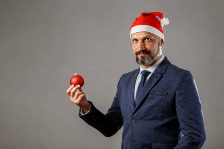 一个留着胡子的男人穿着蓝色夹克，戴着一顶灰色背景的新年帽，手里拿着一个红色的圣诞舞会送给云杉。年轻圣诞老人的概念。