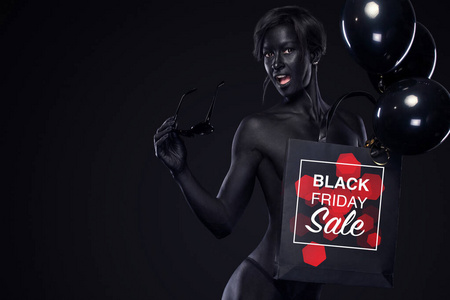 商店的黑色星期五销售概念。带着人体艺术和面部艺术的购物妇女拿着包和气球在黑暗的背景下度假。