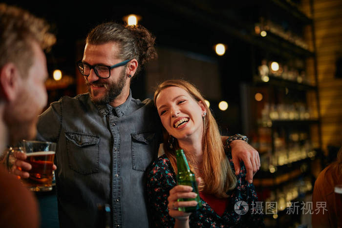 英俊的白人男子戴着眼镜拥抱着他的女朋友,一边和她聊天,一边在酒吧里