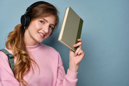 一个穿着粉色运动衫戴着耳机的女孩站在蓝色的背景上，看着画框，举着一本书。