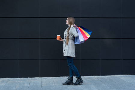 快乐的年轻女人拿着五颜六色的袋子和纸杯。背景为黑色街道墙