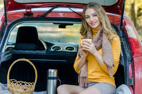 一个漂亮的金发女孩坐在一辆红色汽车的后备箱里，微笑着，