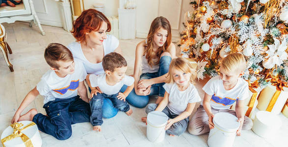 平安夜，快乐的家庭母亲和五个孩子在圣诞树旁放松地玩耍。妈妈，女儿，儿子们在带冬季装饰的灯光房里。新年庆祝横幅
