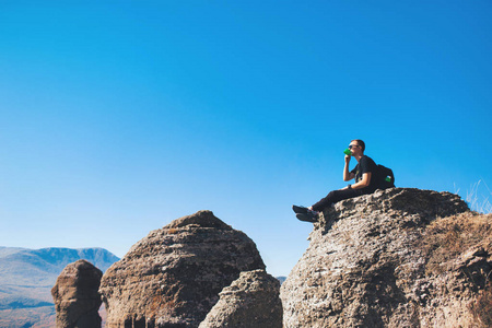 一个背着背包的家伙坐在山上的岩石上，喝茶，欣赏大自然的景色。