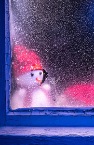 悲伤的雪人透过窗户看。窗外还以冰雪为背景，为您装饰冬季森林雪人景观。圣诞节和晴朗寒冷的日子。