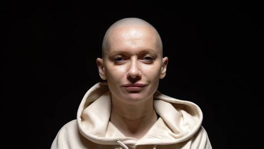 化疗帽头戴连帽衫的光头女人看着镜头,悲伤地笑了起来.