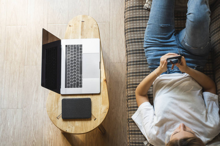 女人躺在沙发上用智能手机，桌上放着笔记本电脑和电子书阅读器。在线学习，自由职业者。自雇或自由职业的女孩使用电话，下班休息时随身携