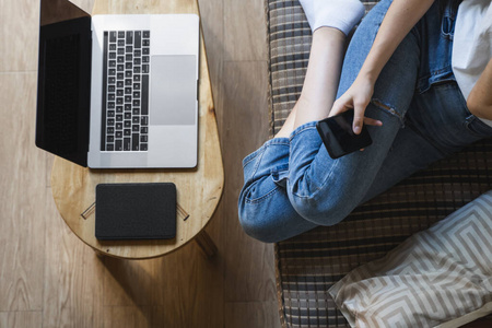 一位女士拿着笔记本电脑坐在沙发上用智能手机。在线学习和工作，自由职业者。一个个体户女孩坐在沙发上，桌上放着电话和电子书阅读器，手