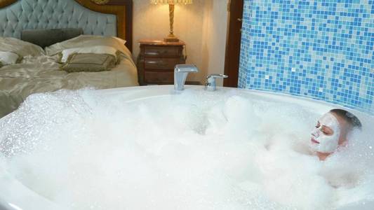 一个秃顶的年轻女子,脸上戴着面具,躺在泡沫浴缸里,她微笑着吹着泡沫