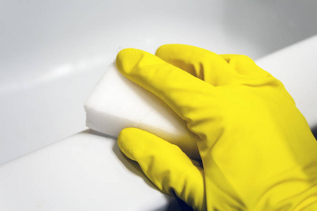 一只戴着黄色橡胶手套的手用三聚氰胺海绵擦拭白色浴缸的表面。在现代化的浴室里打扫卫生。选择性聚焦。特写视图