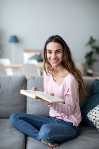 Woman with a book on sofa looking up at the camera with a smile.