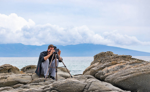 年轻英俊的男摄影师坐在海边的岩石上安装手机三脚架.