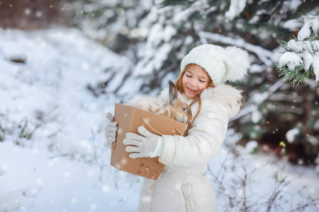 雪林里，一个穿着白色蓬松羽绒服的女孩把一只装饰性的活兔放在盒子里