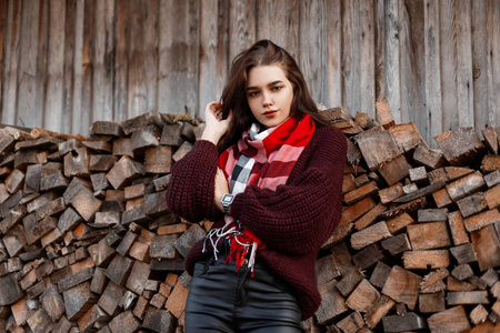 可爱的农村年轻女子穿着时髦的针织毛衣，戴着时髦的围巾，站在一堵用原木砌成的旧木墙上。一个美丽的女孩在温暖的秋日里在村里休息。