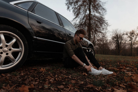 穿着白色运动鞋,戴着墨镜,正在城外一辆现代黑色汽车附近休息