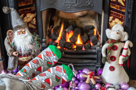 穿着圣诞袜的脚在壁炉旁。女人在温暖的炉火旁放松，穿着滑稽的袜子暖脚。双脚靠近。冬季和圣诞节概念